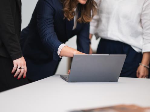 Hand zeigt auf einen Laptop, Personen stehen daneben.