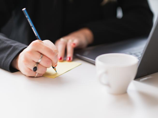 Mano scrive su un post-it accanto a un laptop.